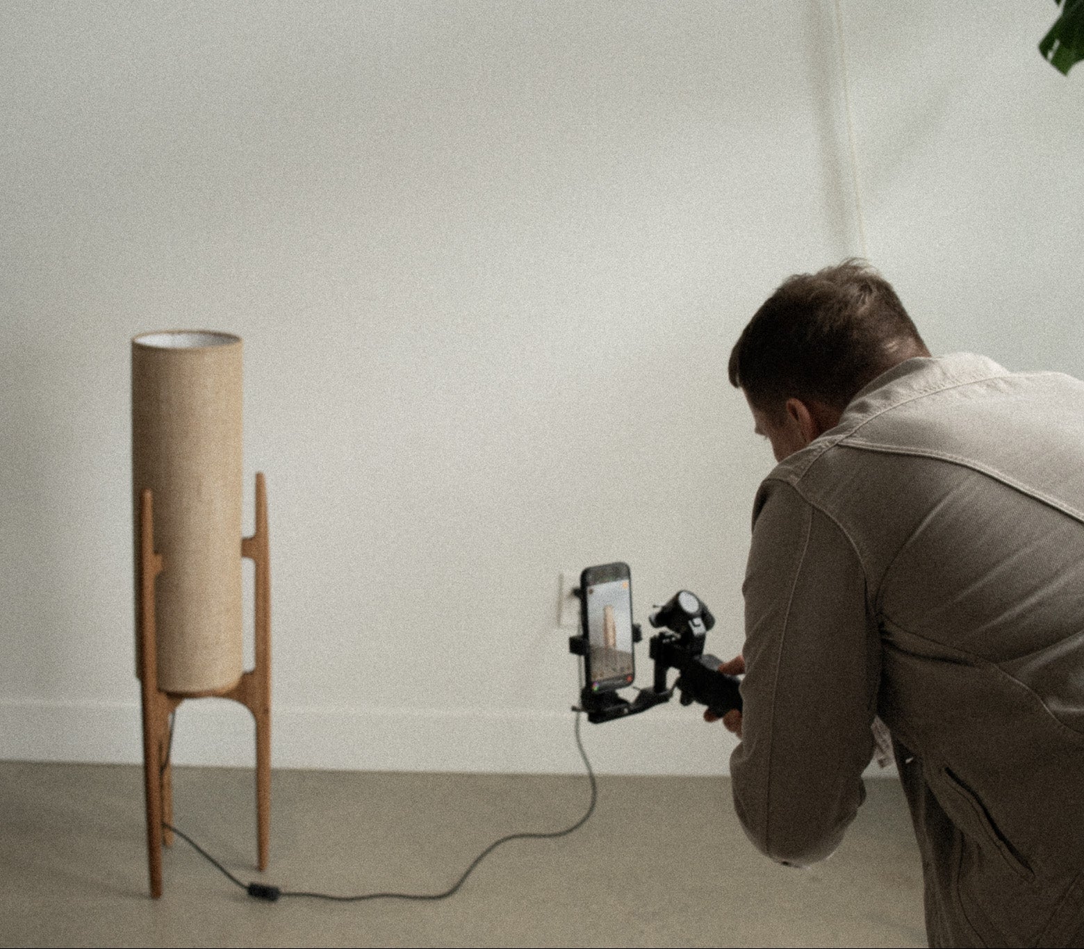 Person using a smartphone with a gimbal to photograph a lamp in a minimalistic room.