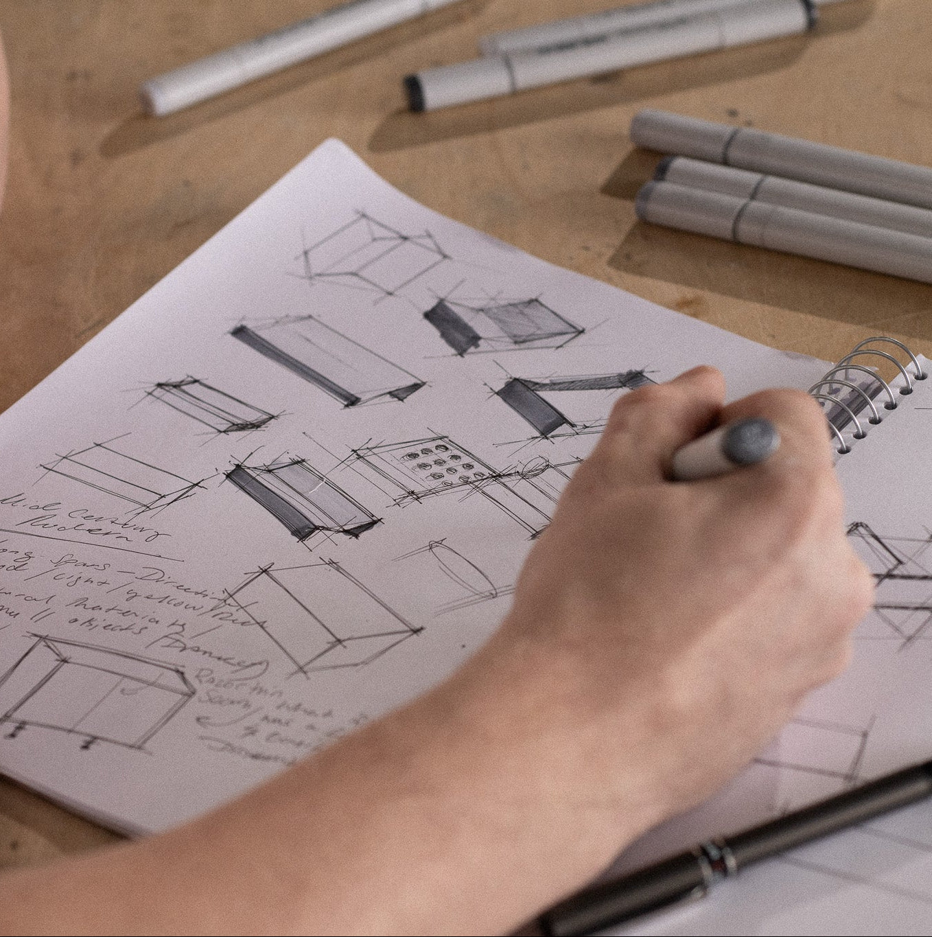 Person sketching furniture designs on a piece of paper with markers on a wooden table.